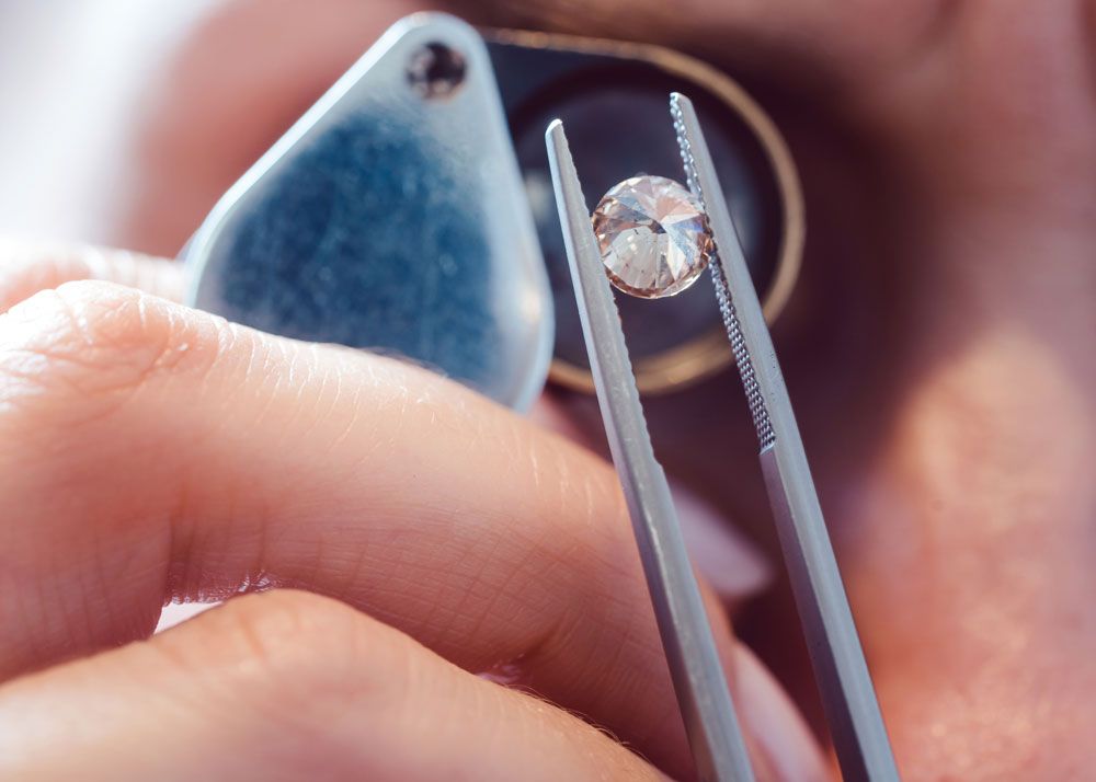 A closeup of a jeweler's right eye,  behind a loupe, inspecting a diamond held by tweezers at close magnification.