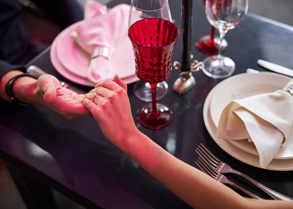 A man and woman holding hands at a table in a restaurant. The woman is wearing a diamond ring and it appears to be an engagement dinner.